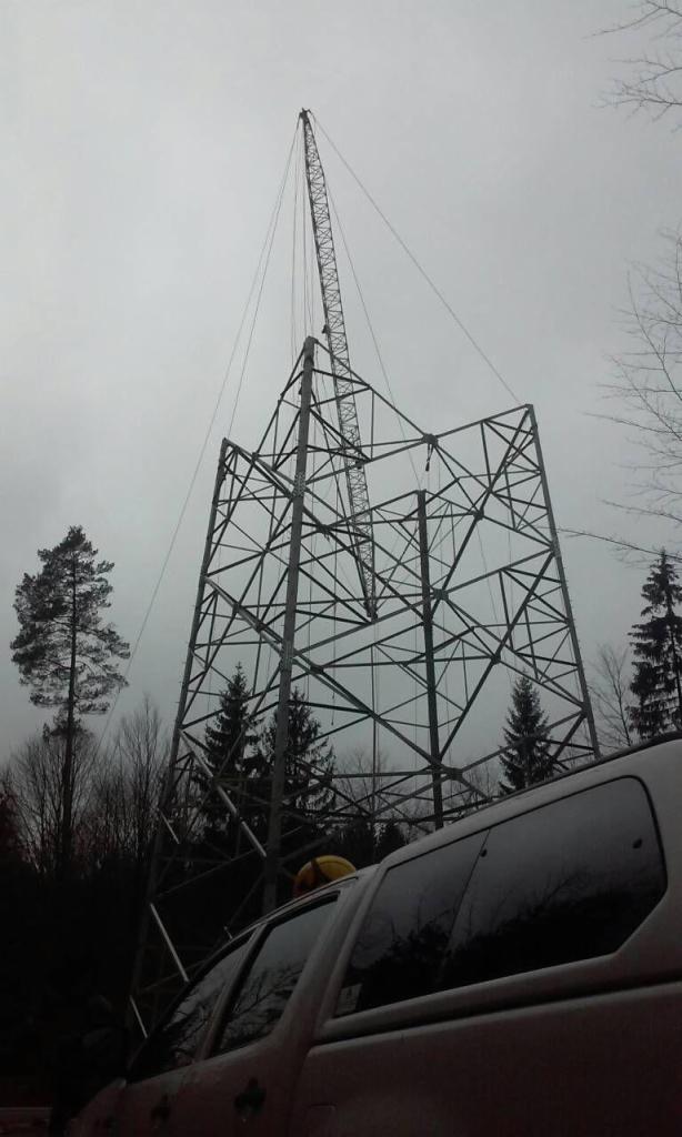 Montaż metalowej konstrukcji wieży energetycznej przy użyciu dźwigu na tle pochmurnego nieba i drzew, częściowo zasłoniętej przez białe auto z żółtym elementem na dachu.
