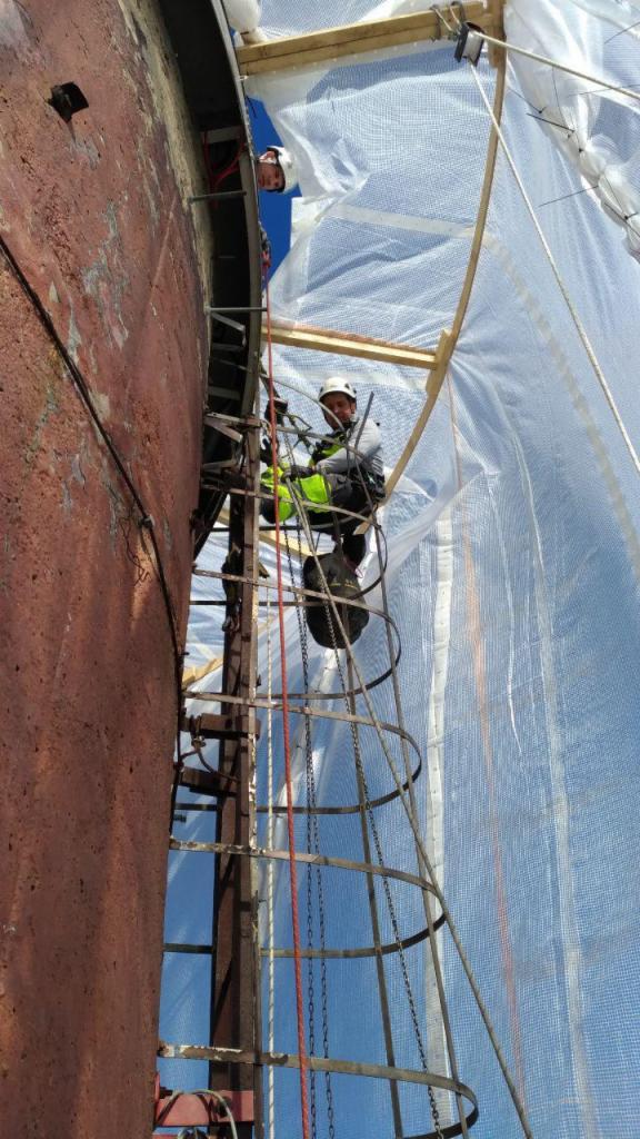 Pracownik na metalowej drabinie przymocowanej do dużej, rdzewiejącej konstrukcji, prawdopodobnie zbiornika, zabezpieczonej przezroczystą folią i drewnianymi rusztowaniami, drugi pracownik widoczny...