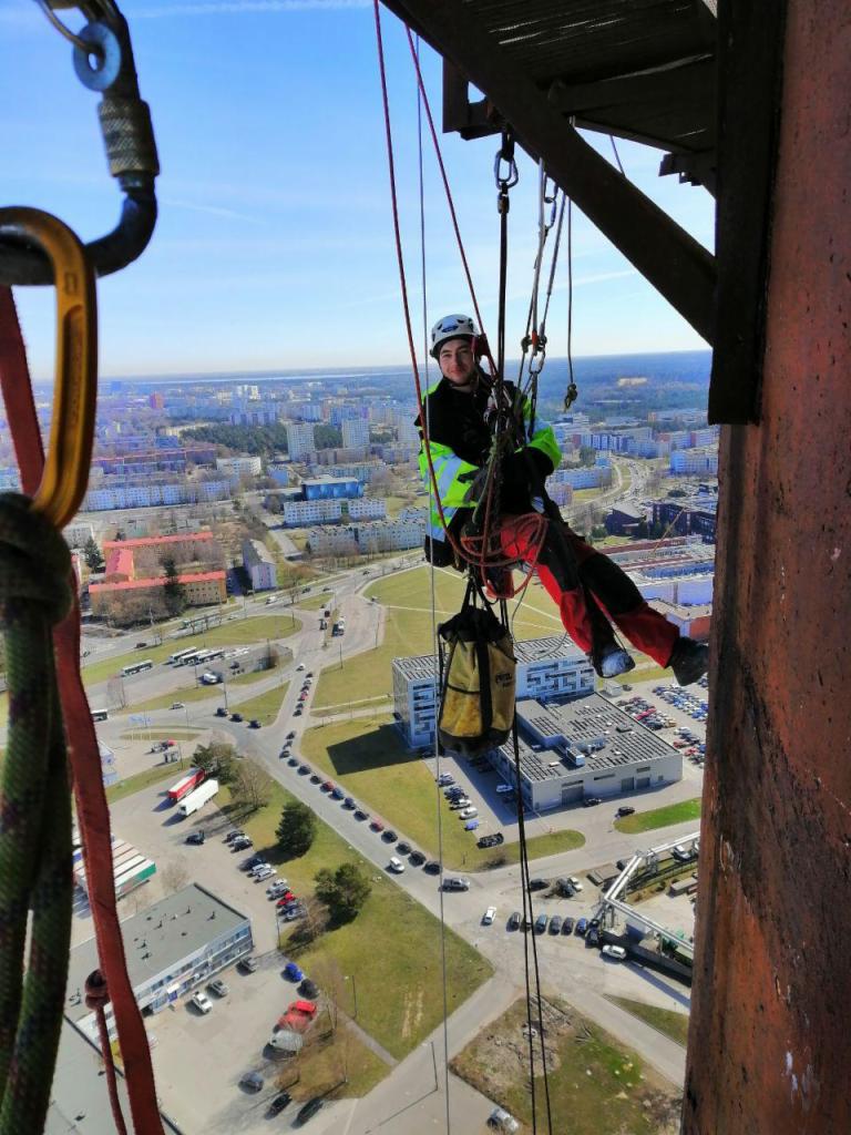 Pracownik na wysokościach, w kasku i uprzęży, zwisa na linach z metalowej konstrukcji, prawdopodobnie masztu; widok na miasto w tle.