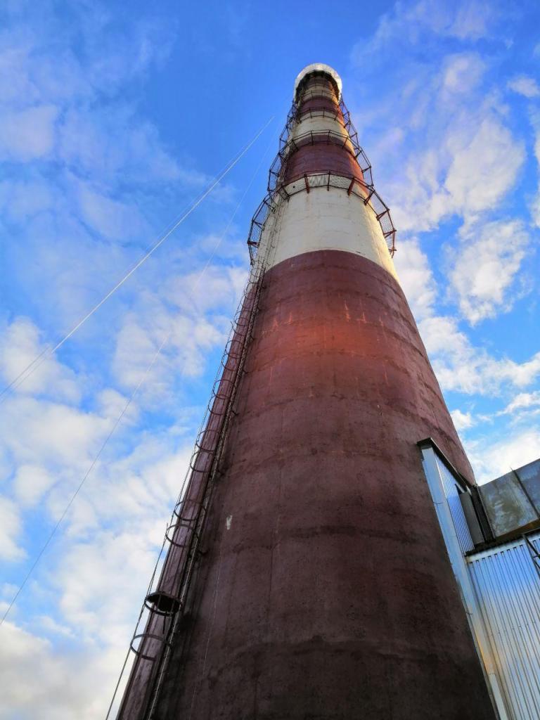 Wysoki komin przemysłowy z metalową drabiną na tle błękitnego, pochmurnego nieba, z widocznymi śladami malowania w kolorach białym i ceglastym.