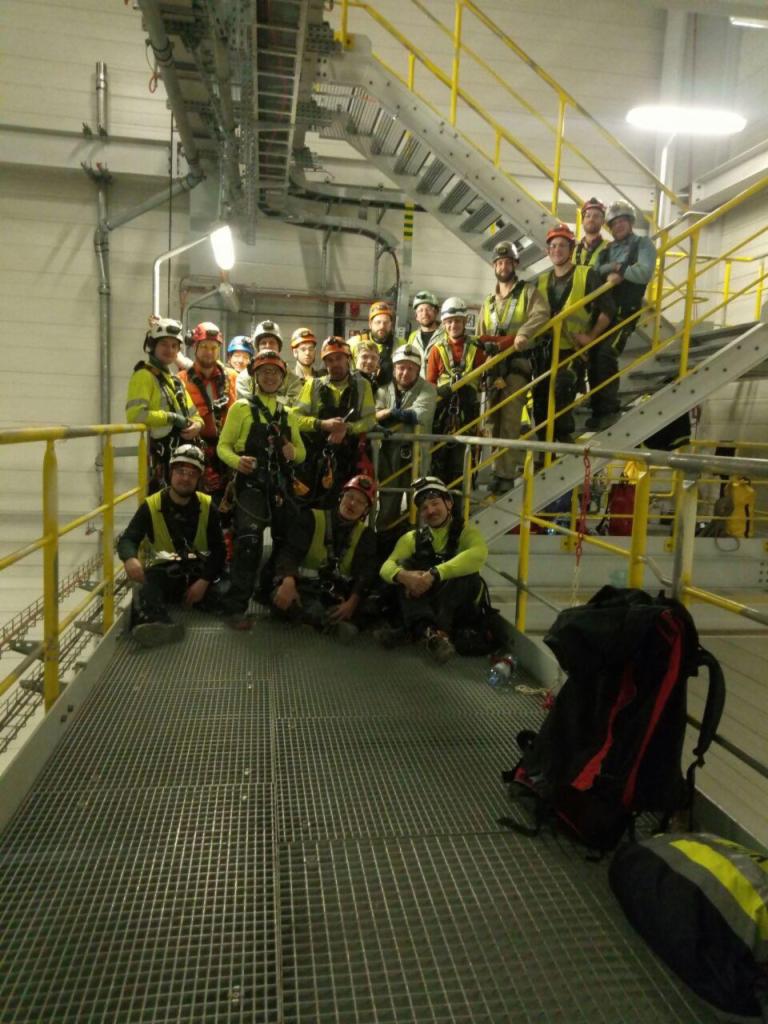 Grupa pracowników w kaskach i uprzężach na metalowej konstrukcji w hali przemysłowej, pozujących do zdjęcia po zakończonych pracach wysokościowych.