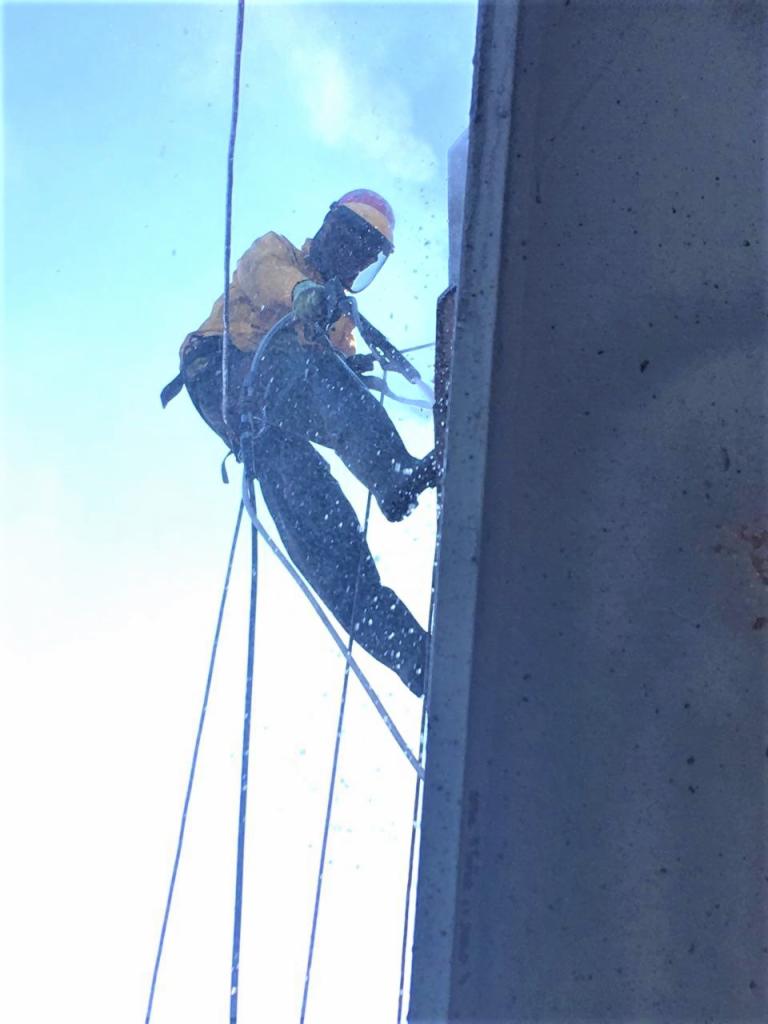 Pracownik w uprzęży, z widoczną osłoną twarzy, wykonuje prace na wysokości na betonowej konstrukcji. Z dołu widoczne liny i niebo.