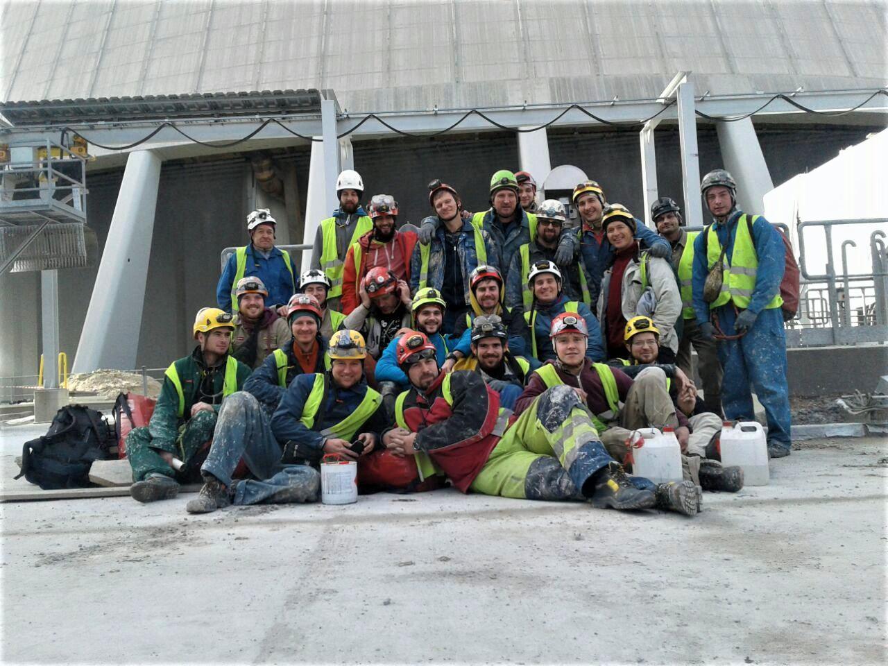 Duża grupa uśmiechniętych pracowników budowlanych w kaskach i odblaskowych kamizelkach pozuje na tle betonowej konstrukcji chłodni kominowej, z widocznymi pojemnikami z farbą u ich stóp.