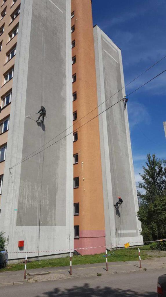 Trzech alpinistów przemysłowych odświeża elewację wysokiego bloku mieszkalnego, używając lin i sprzętu do pracy na wysokościach. Widoczne ślady zabrudzeń na szarej ścianie budynku.