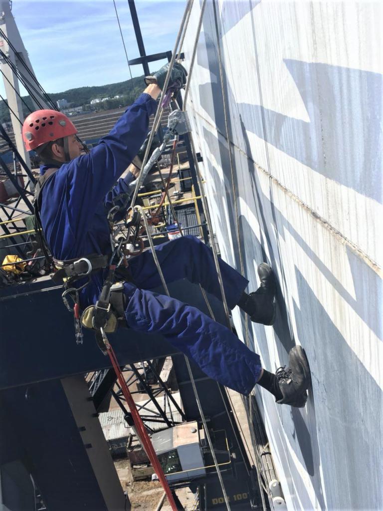 Pracownik w kombinezonie i kasku, zabezpieczony linami, maluje zewnętrzną ścianę zbiornika przemysłowego na wysokości, z widocznymi zaciekami świeżej farby.
