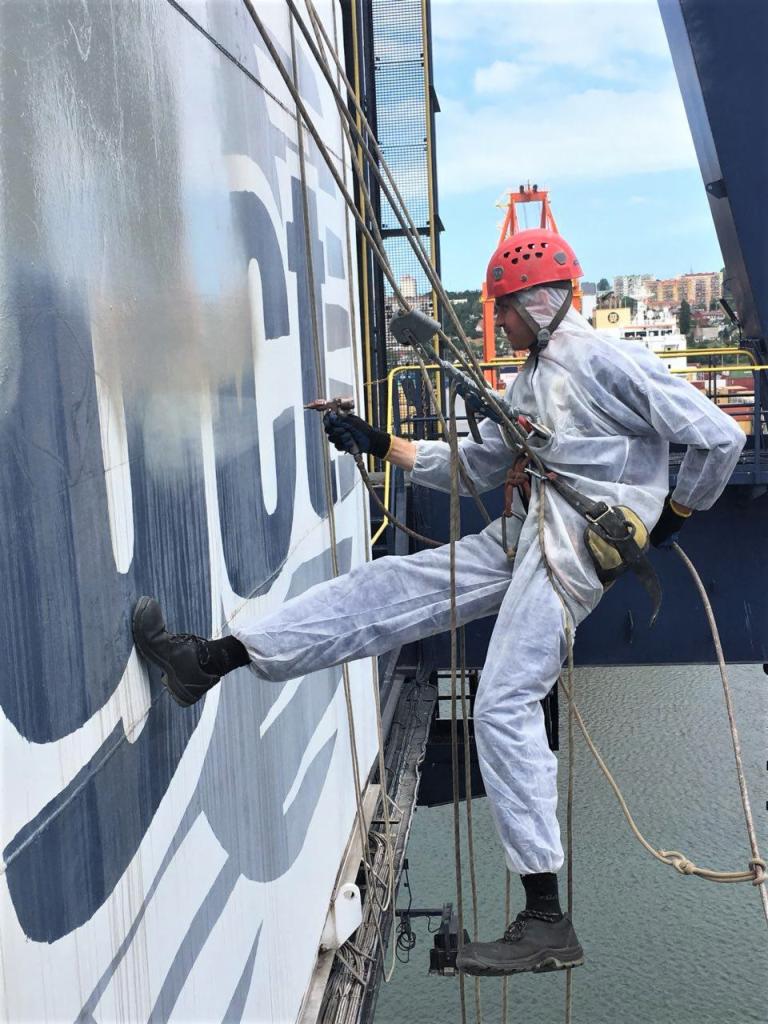 Pracownik w kombinezonie ochronnym i czerwonym kasku, zawieszony na linach, natryskowo nanosi szarą farbę na dużą powierzchnię kontenera z częściowo widocznym, białym logotypem; w tle widać miasto...