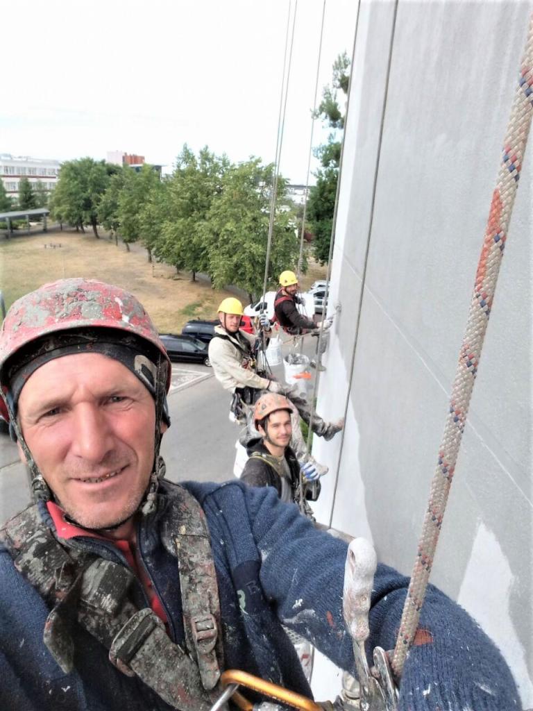 Trzech alpinistów zabezpieczonych linami, w hełmach i ubraniach roboczych z widocznymi śladami farby, wykonuje prace na wysokościach, malując zewnętrzną ścianę budynku.