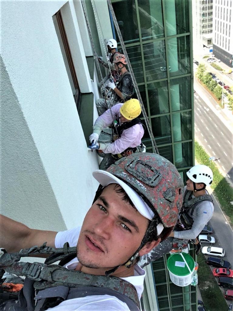 Ekipa alpinistów podczas prac na wysokościach, zabezpieczona linami, maluje elewację budynku. Jeden z pracowników, z kaskiem pomalowanym farbą, robi selfie na tle miasta.