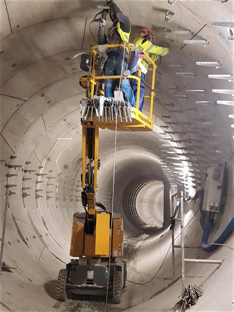 Dwóch pracowników na podnośniku koszowym montuje elementy wewnątrz betonowego tunelu. Widoczne liczne uchwyty mocujące na ścianach tunelu i narzędzia na platformie podnośnika.