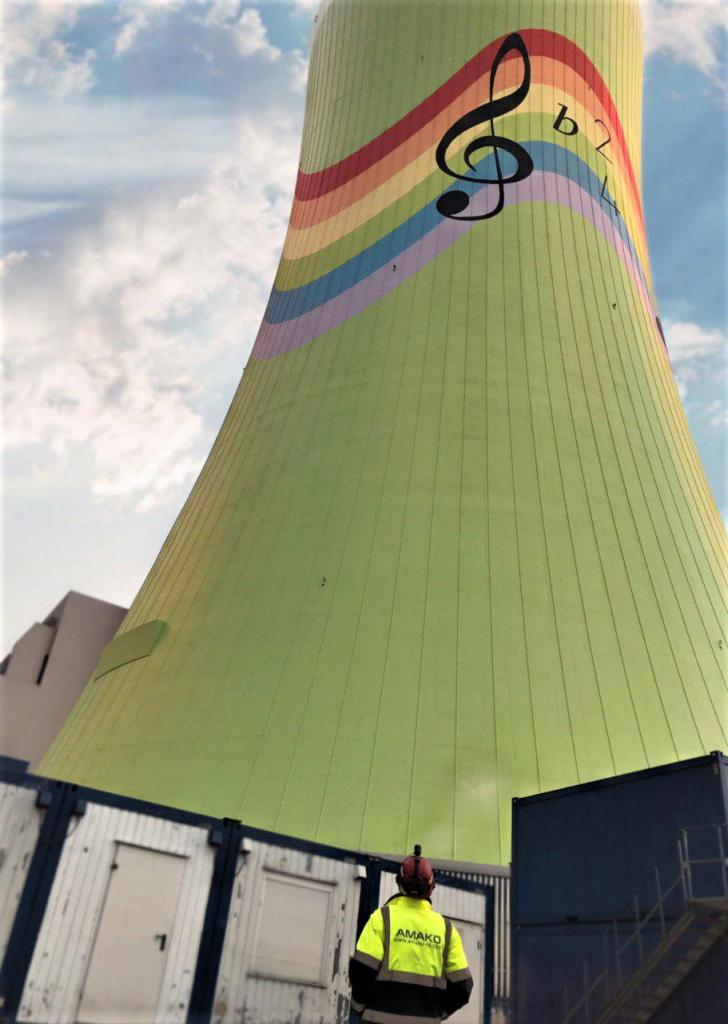 Wysoka wieża chłodnicza pomalowana w pastelowe kolory tęczy z motywem muzycznym (klucz wiolinowy i bemole), pracownik w odblaskowej kurtce firmy AMAKO na tle kontenerów biurowych.