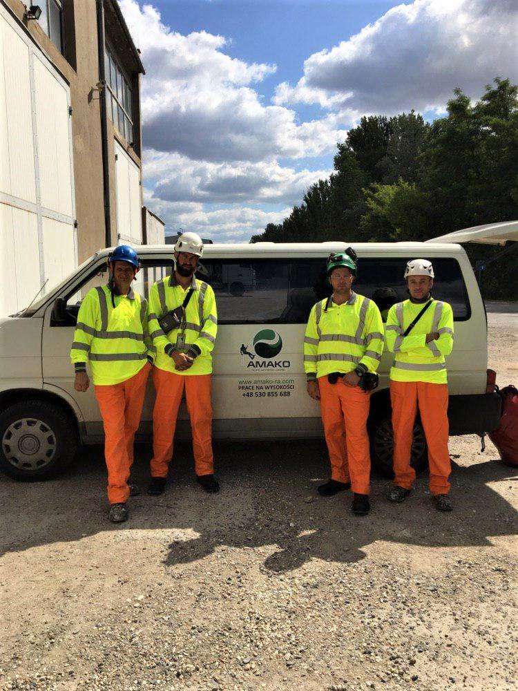 Czterech pracowników w odblaskowych strojach i kaskach stoi przed vanem firmy AMAKO na żwirowym placu, z budynkiem i drzewami w tle.
