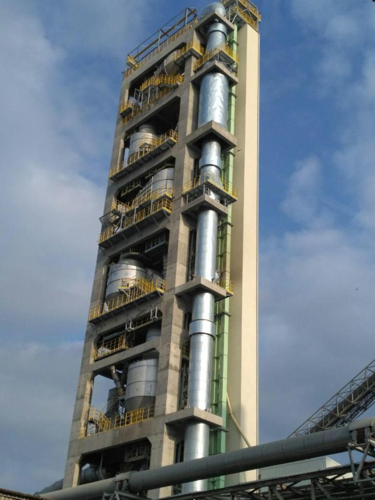 Wysoka, przemysłowa konstrukcja z betonu i metalu, z widocznymi rurami i zbiornikami, zabezpieczona żółtymi barierkami, na tle błękitnego nieba z chmurami.