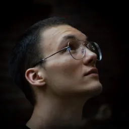 Młody mężczyzna w okularach spogląda w górę, oświetlony z dołu, na ciemnym tle. Widoczny profil twarzy i cienkie oprawki okularów.