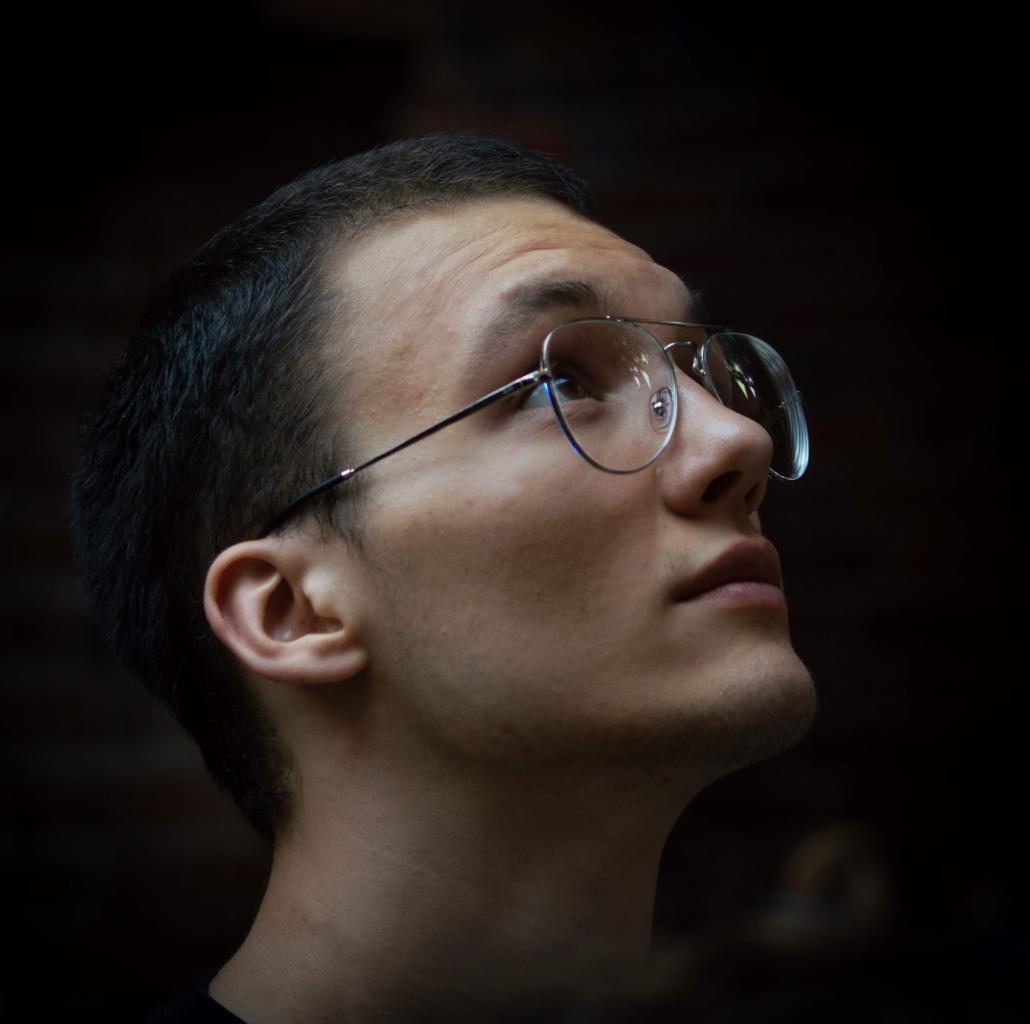 Młody mężczyzna w okularach spogląda w górę, oświetlony z dołu, na ciemnym tle. Widoczny profil twarzy i cienkie oprawki okularów.