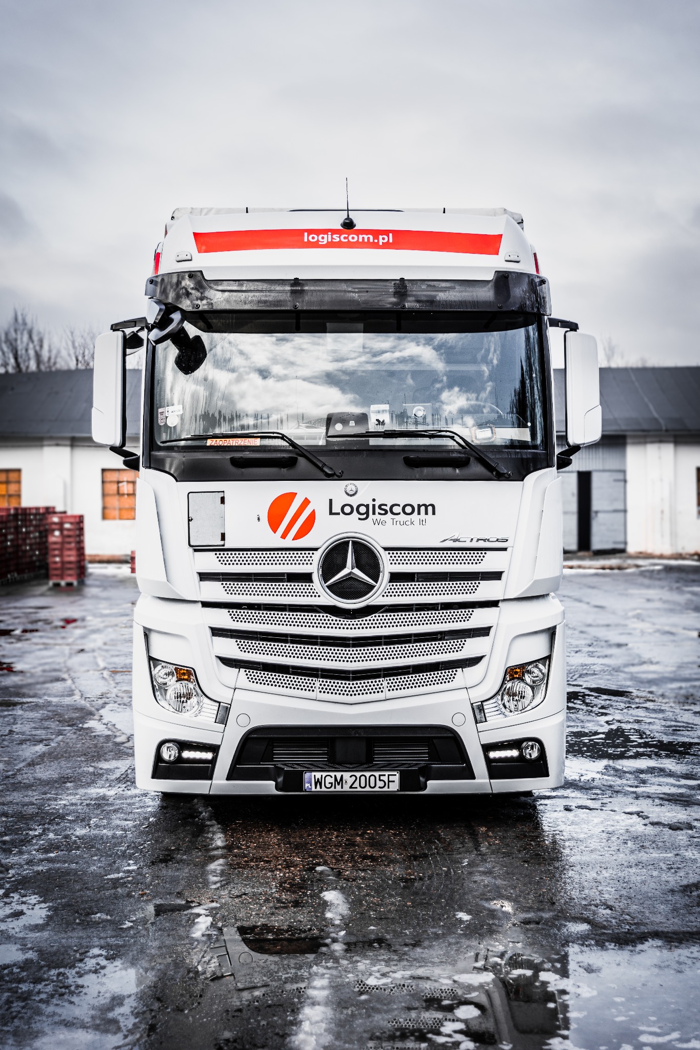 Przód białej ciężarówki marki Mercedes z logo Logiscom i numerem rejestracyjnym WGM 2005F, stojącej na mokrym placu przed budynkiem magazynowym w pochmurny dzień.