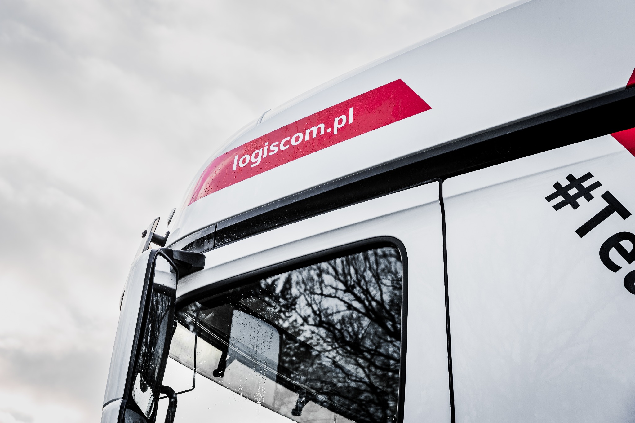 Fragment kabiny białej ciężarówki z czerwonym logo logiscom.pl i hasztagiem #Te, widok z dołu na tle pochmurnego nieba, krople deszczu na szybie.