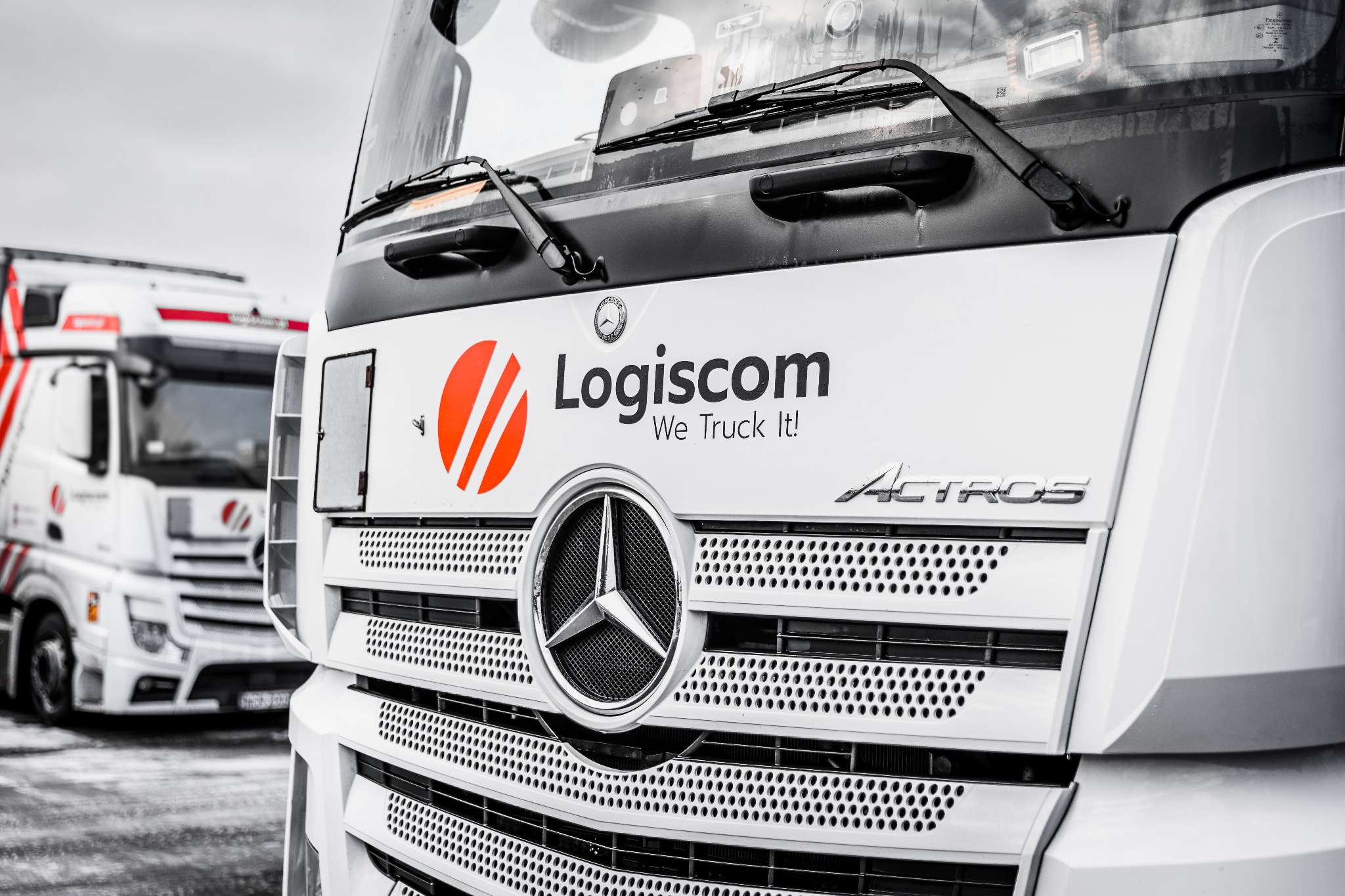Przód białej ciężarówki Mercedes-Benz Actros z logo firmy Logiscom na tle innej ciężarówki tej samej firmy.