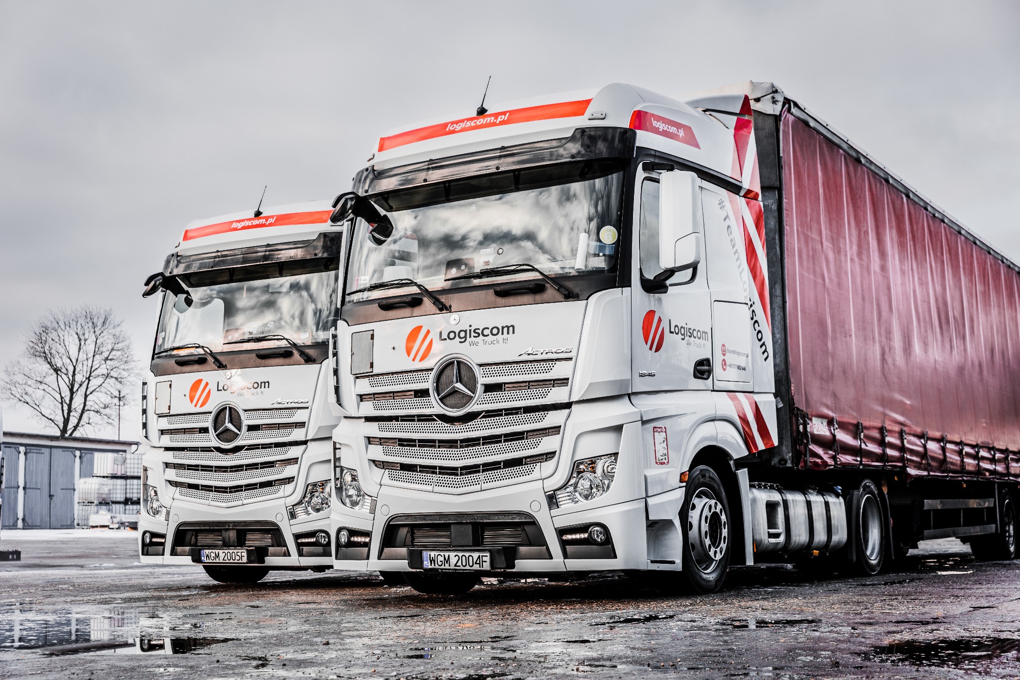 Dwa białe ciągniki siodłowe marki Mercedes-Benz Actros z czerwonymi plandekami naczep, zaparkowane na mokrym placu, z logo Logiscom i hasłem 'We Truck It!'