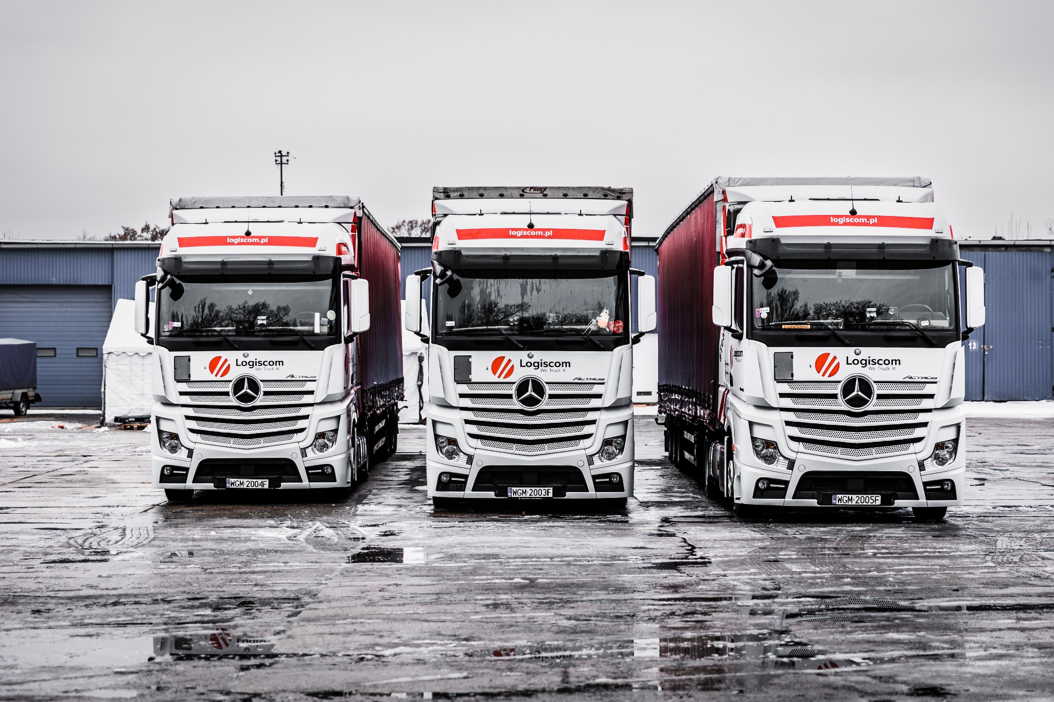 Trzy białe ciężarówki marki Mercedes Actros z logo Logiscom ustawione równolegle na mokrym placu, widoczne tablice rejestracyjne WGM 2004F, WGM 2003F i WGM 2005F, w tle budynek magazynowy.