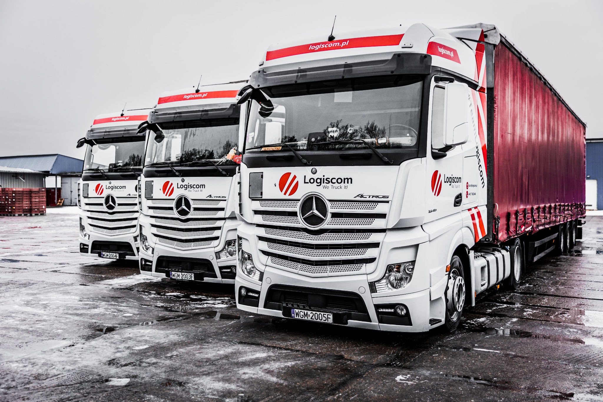 Trzy białe ciężarówki Mercedes Actros z logo Logiscom ustawione w rzędzie na mokrym placu, widoczna czerwona naczepa z tyłu.
