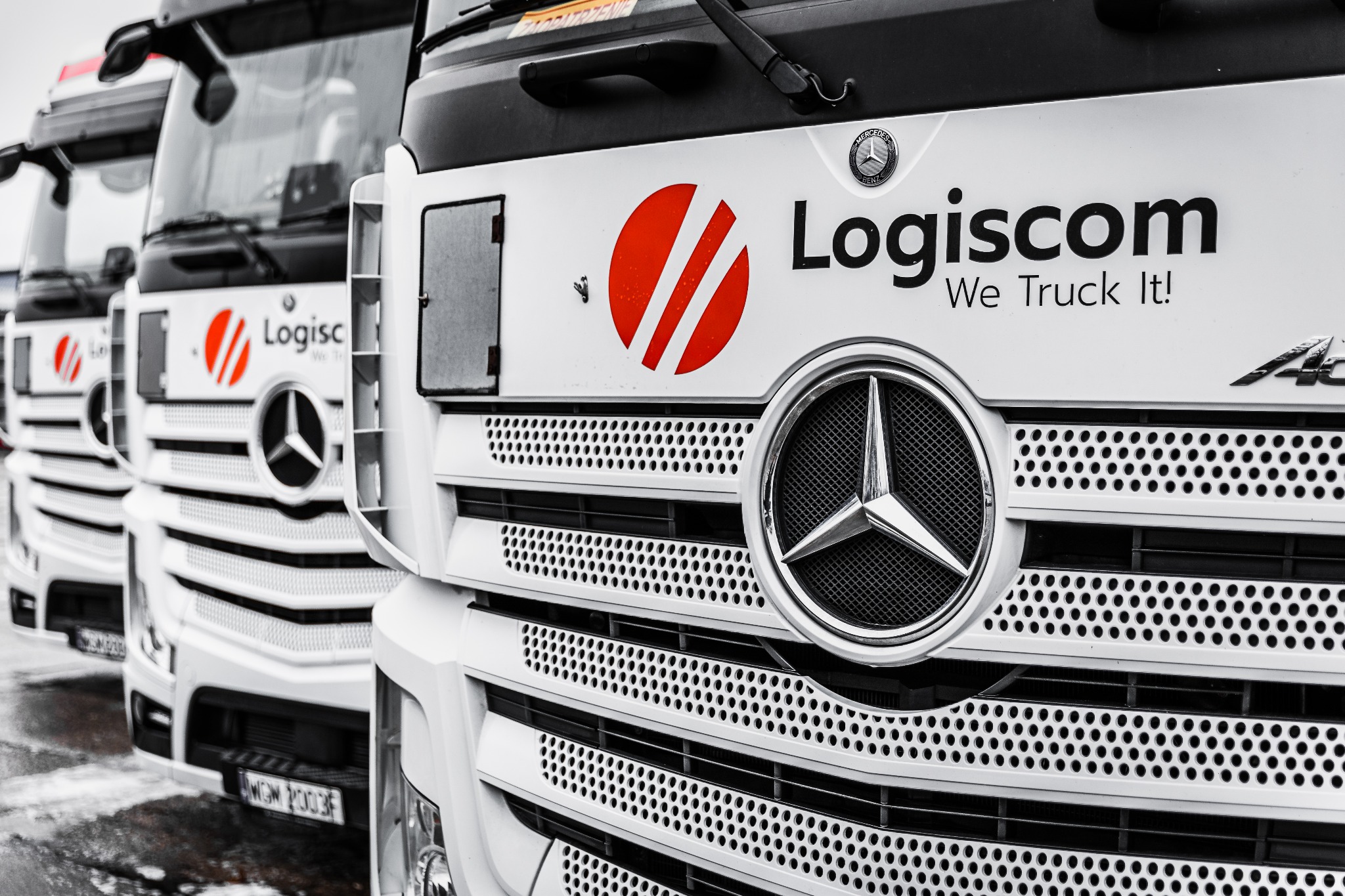 Flota białych ciężarówek marki Mercedes z logo firmy Logiscom, zaparkowanych w rzędzie, widok z przodu na grille i logotypy.