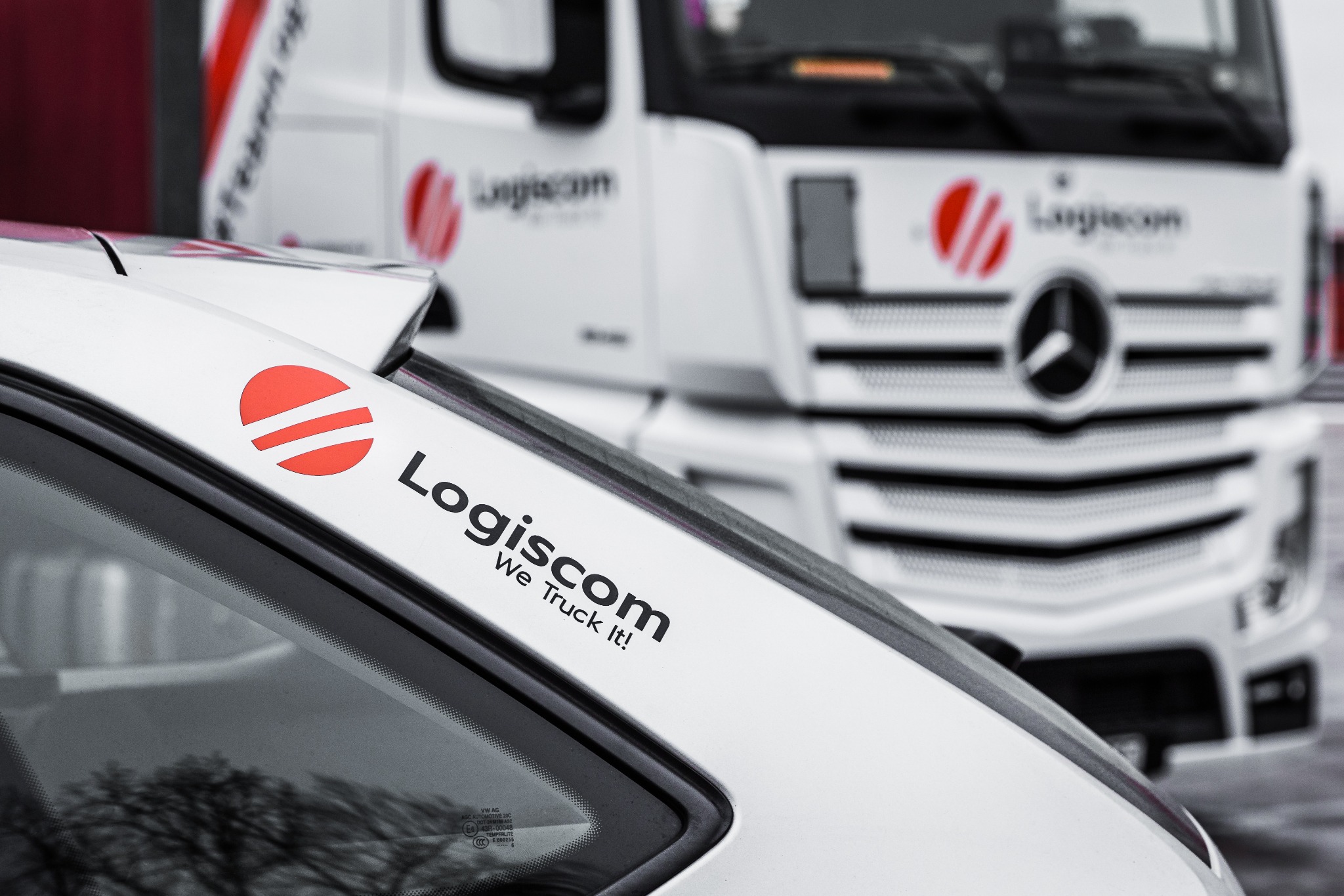 Biała kabina samochodu ciężarowego z logo Logiscom i napisem 'We Truck It!' na tle drugiego, rozmytego samochodu ciężarowego tej samej firmy.