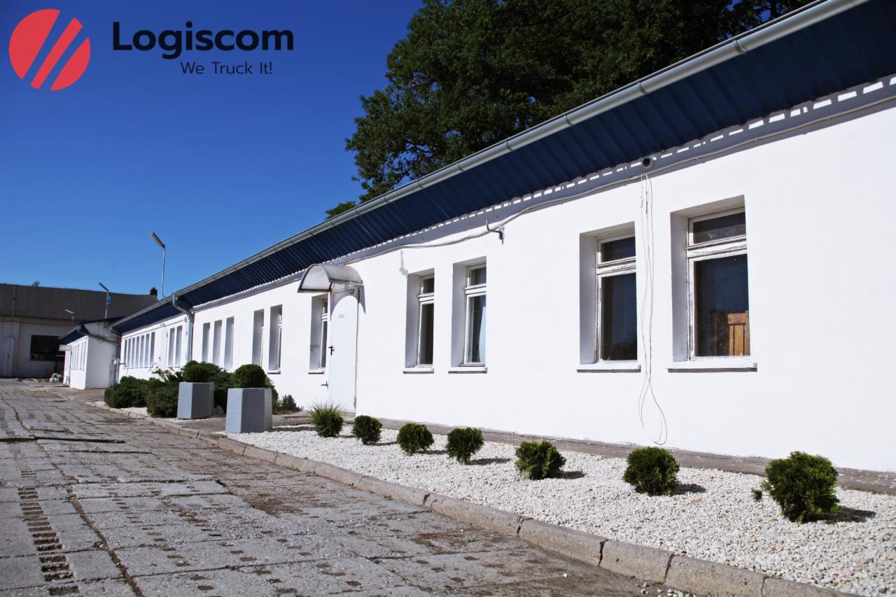 Biały budynek magazynowy z niebieskim dachem pod błękitnym niebem, logo firmy Logiscom w lewym górnym rogu.