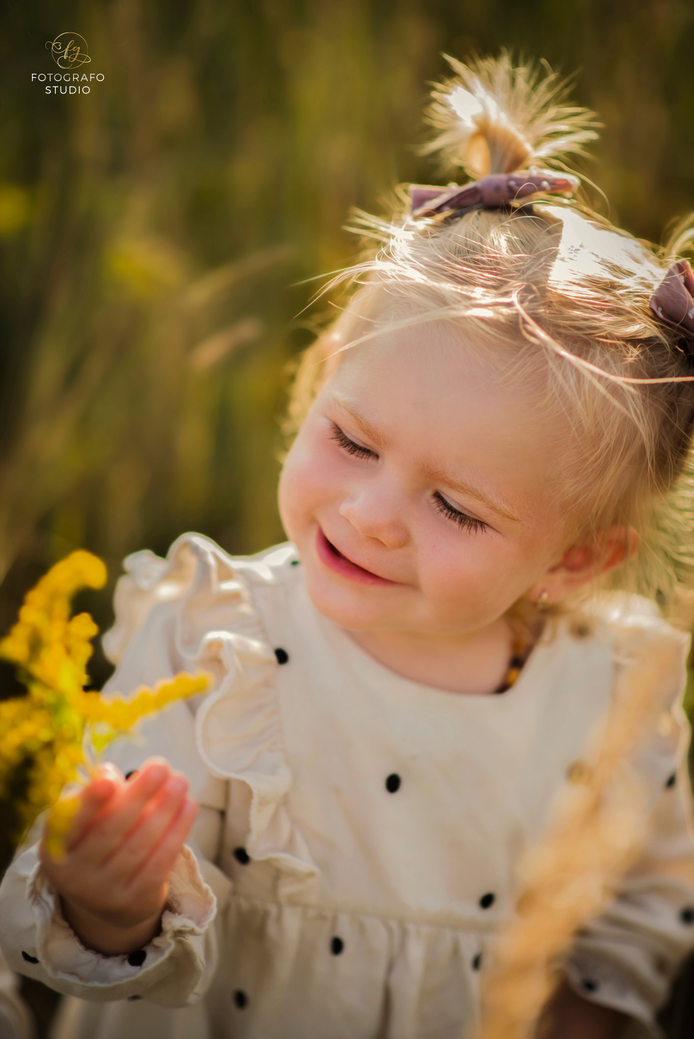 Uśmiechnięta dziewczynka z blond włosami i kokardami, trzymająca żółte kwiaty na łące, w sukience w czarne kropki.