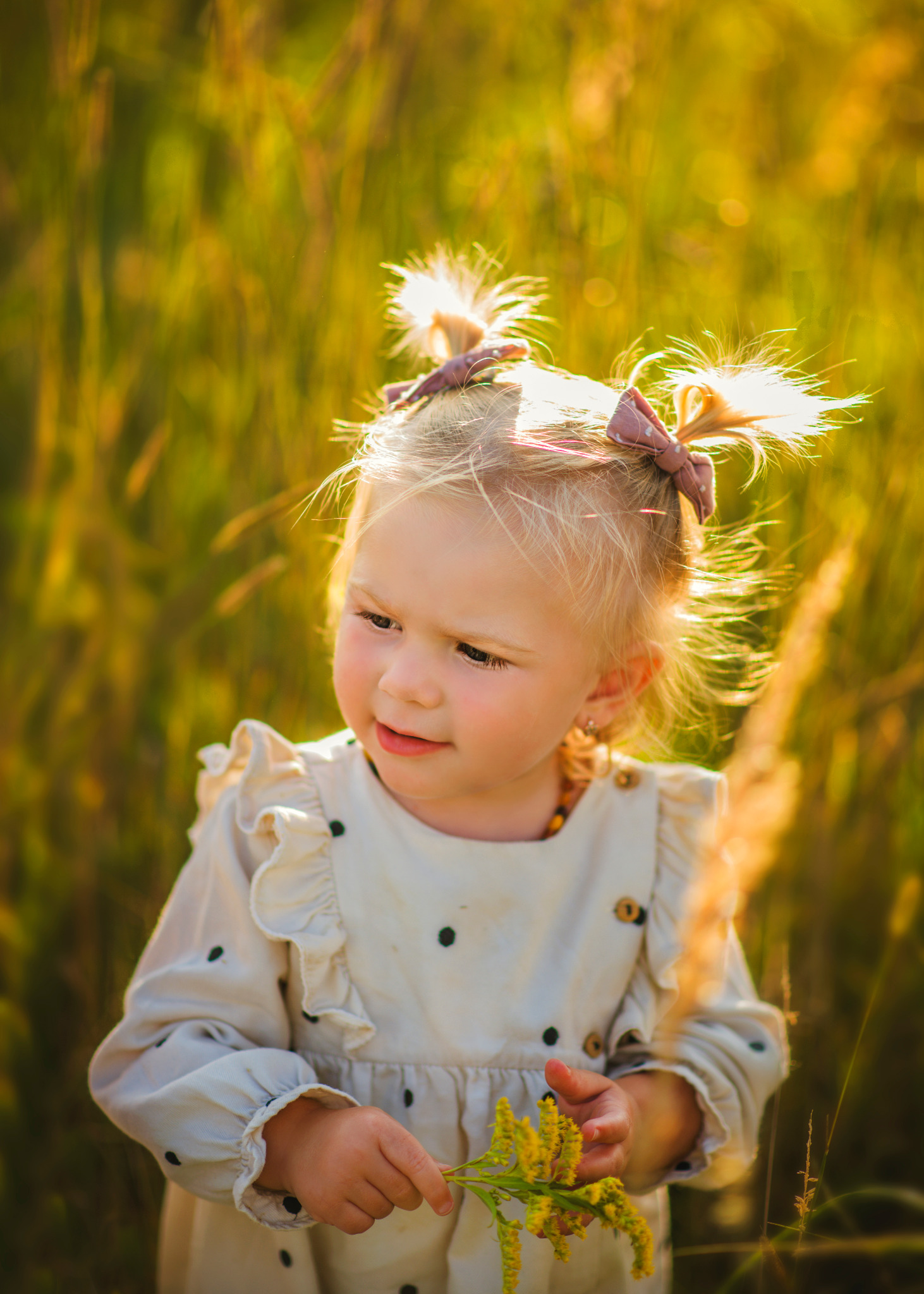 Dziewczynka z blond włosami spiętymi w dwa kucyki z brązowymi kokardkami, w sukience w czarne kropki, trzymająca żółte kwiaty na tle rozświetlonej słońcem trawy.