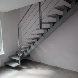 Schody wspornikowe z drewnianymi stopniami i metalową konstrukcją nośną oraz minimalistyczną balustradą z cienkich prętów, zamontowane w jasnym wnętrzu z panelami podłogowymi.