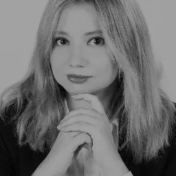 Portret kobiety w czarno-białej tonacji, z dłońmi splecionymi pod brodą, ubrana w białą koszulę i czarną marynarkę, z delikatnym uśmiechem.