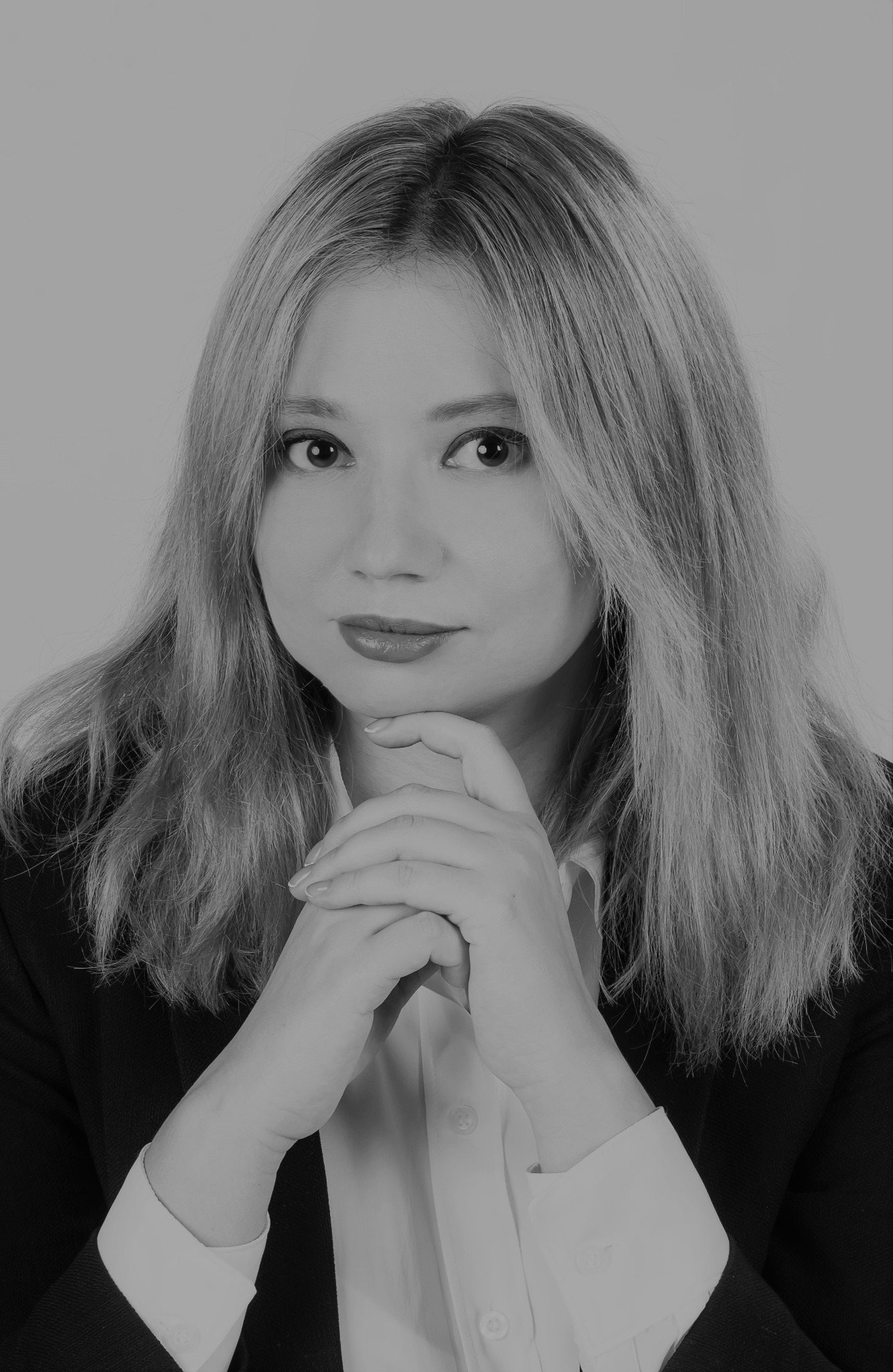 Portret kobiety w czarno-białej tonacji, z dłońmi splecionymi pod brodą, ubrana w białą koszulę i czarną marynarkę, z delikatnym uśmiechem.