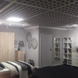 Jasne wnętrze sypialni w stylu skandynawskim z drewnianą podłogą, łóżkiem z szaro-białą pościelą, szafą z jasnego drewna i ażurowym sufitem podwieszanym.