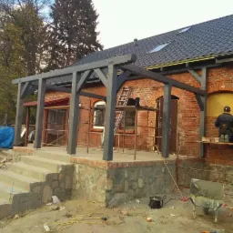 Budowa zadaszenia tarasu z drewnianą konstrukcją przy ceglanym domu. Widoczne schody wejściowe z kamienną podmurówką, rusztowania, narzędzia i murarz przy pracy.