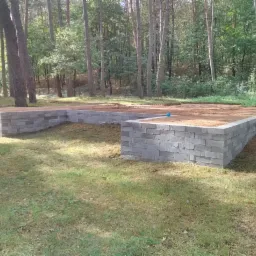 Wykop fundamentowy z bloczków betonowych, wypełniony piaskiem, widoczny na tle lasu sosnowego w słoneczny dzień. Częściowo wykoszona trawa wokół fundamentu.