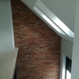 Ściana z cegły klinkierowej na poddaszu, widok na okno dachowe i podłogę z narzędziami, surowe wykończenie.