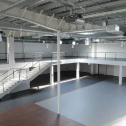 Przestronne wnętrze hali z antresolą i schodami, widok z góry, oświetlenie przemysłowe, surowy beton i stalowe elementy konstrukcyjne.