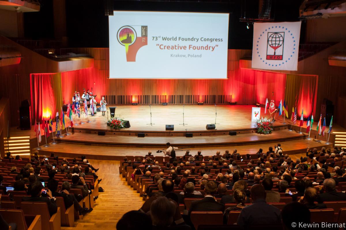 Szeroki kadr na salę kongresową podczas 73. Światowego Kongresu Odlewnictwa w Krakowie, z występem zespołu ludowego na scenie oświetlonej czerwonymi światłami, flagami i ekranem z logo kongresu.