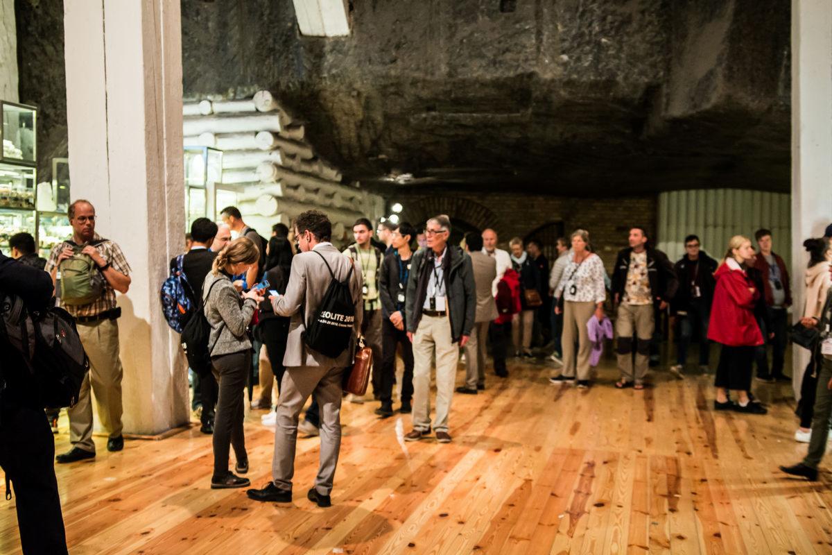 Grupa ludzi w podziemnej sali z drewnianą podłogą i kamiennym stropem, prawdopodobnie podczas zwiedzania lub wydarzenia zorganizowanego w kopalni.