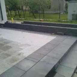 Częściowo ułożona, jasna kostka brukowa na tarasie z niską, kamienną ścianką i schodami. Widoczne narzędzie pracy, jasne tło z zielenią.