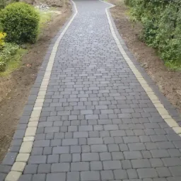 Nowa, szara kostka brukowa z jasnymi obrzeżami prowadząca do garażu z czerwonym autem w tle. Widok z perspektywy pieszego, świeżo ułożona nawierzchnia.