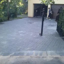 Szara kostka brukowa ułożona na podjeździe do garażu, widoczny fragment ogrodzenia i słupka. Osoba w tle otwiera bramę garażową.