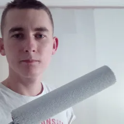 Młody mężczyzna z wałkiem do malowania ścian, widoczna częściowo pomalowana ściana w tle. Ubrany w białą koszulkę z nadrukiem motocyklowym.