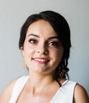 Uśmiechnięta brunetka w białej sukience na tle szarej ściany. Portret kobiety z delikatnym makijażem i złotymi kolczykami, prezentujący profesjonalny wizerunek.