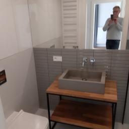 Robert Rybicki - Szara, prostokątna umywalka nablatowa z chromowaną baterią, osadzona na drewnianym blacie z metalowym stelażem w nowoczesnej łazience z szarymi płytkami imitującymi cegłę. W lustrze odbija się...