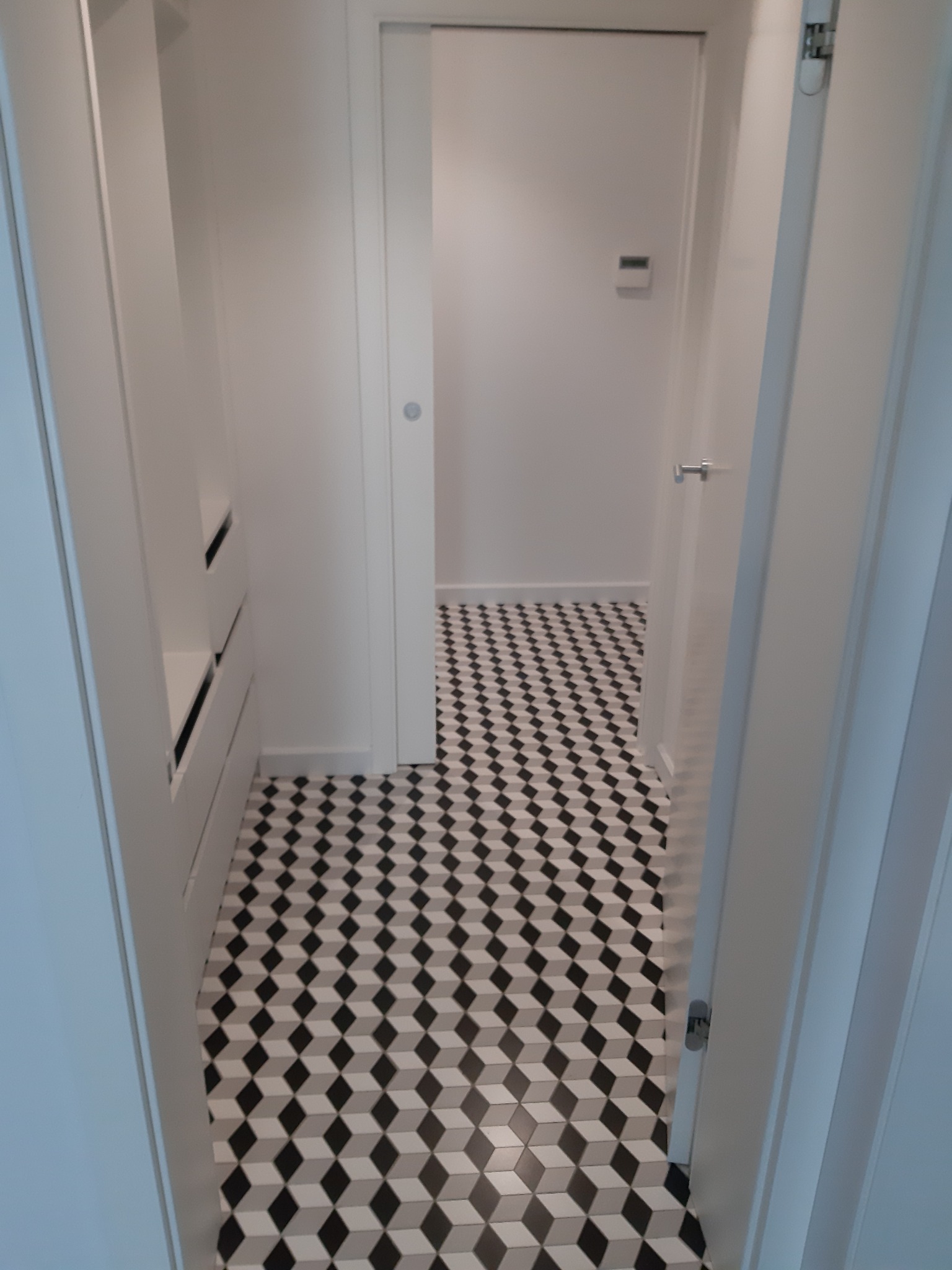 Wnętrze garderoby z białą zabudową meblową i podłogą w czarno-białe geometryczne wzory 3D, widok zza uchylonych drzwi.