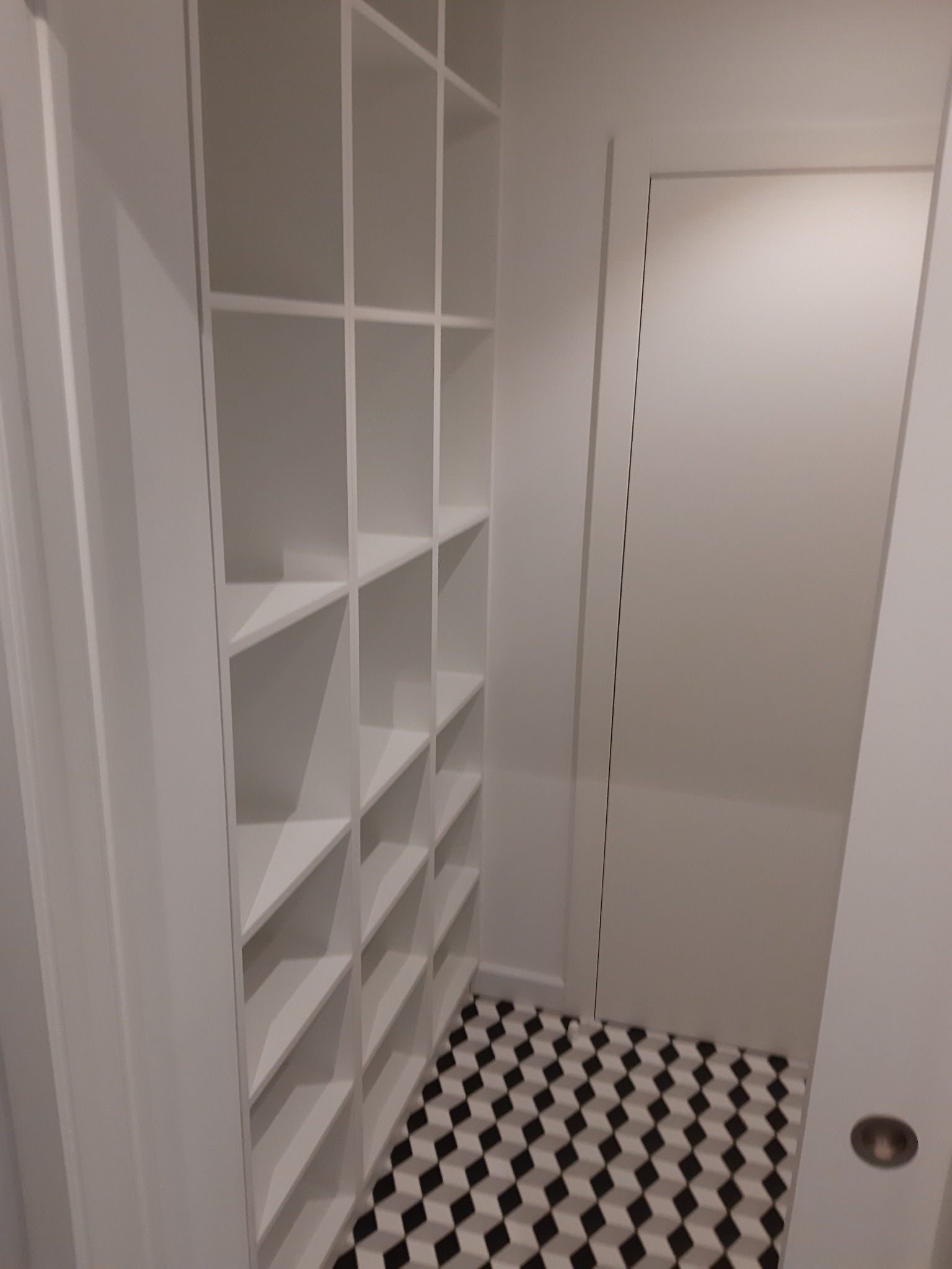 Wnętrze z białą, zabudowaną półką regałową i drzwiami w tle, na podłodze wzór geometryczny czarno-biały.