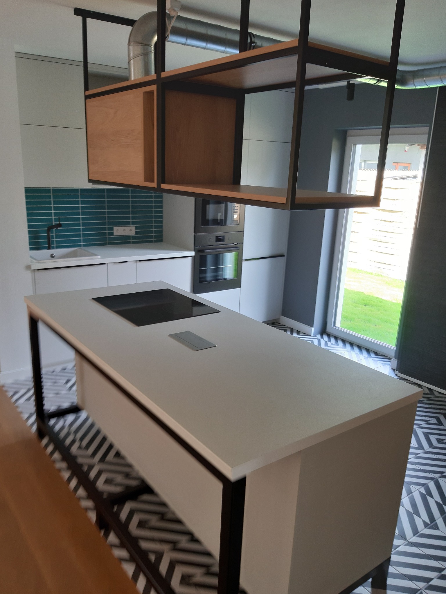 Nowoczesna kuchnia z wyspą kuchenną, wiszącą półką z drewna i metalu, turkusowymi płytkami na ścianie oraz wzorzystą podłogą w biało-czarne zygzaki.