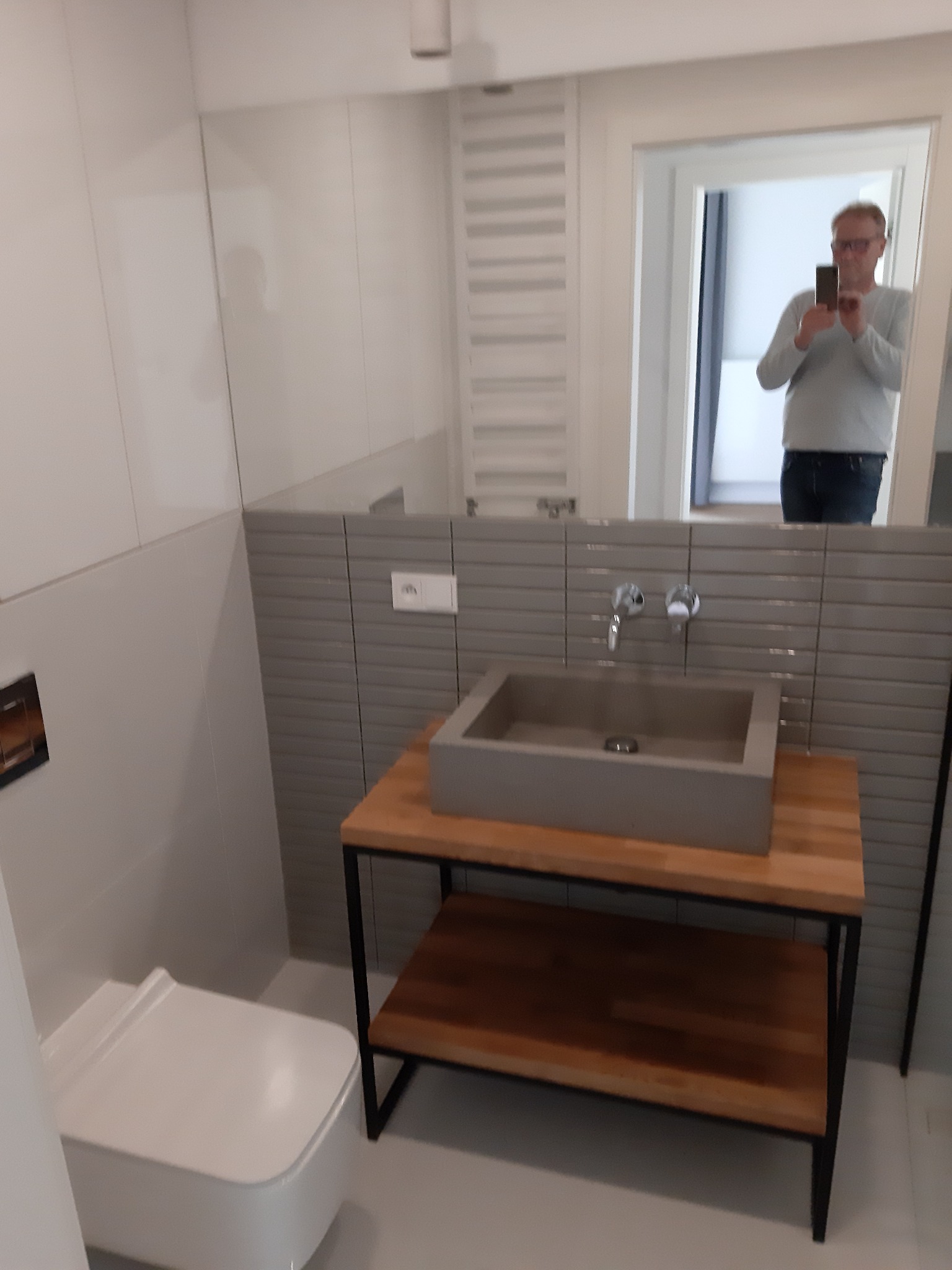 Szara, prostokątna umywalka nablatowa z chromowaną baterią, osadzona na drewnianym blacie z metalowym stelażem w nowoczesnej łazience z szarymi płytkami imitującymi cegłę. W lustrze odbija się...