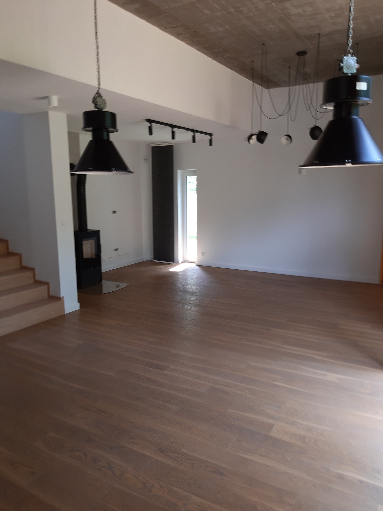 Salon z drewnianą podłogą, betonowym sufitem i industrialnymi lampami wiszącymi, widoczny kominek.